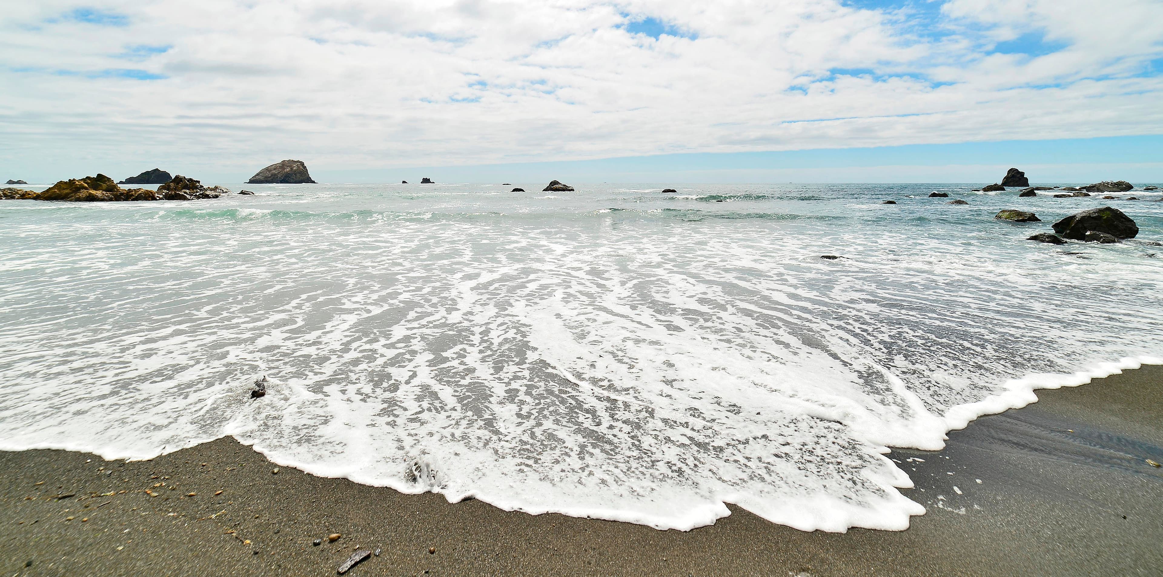 Waves run-up on a beach, and sea stack rocks are seen in the distance.
